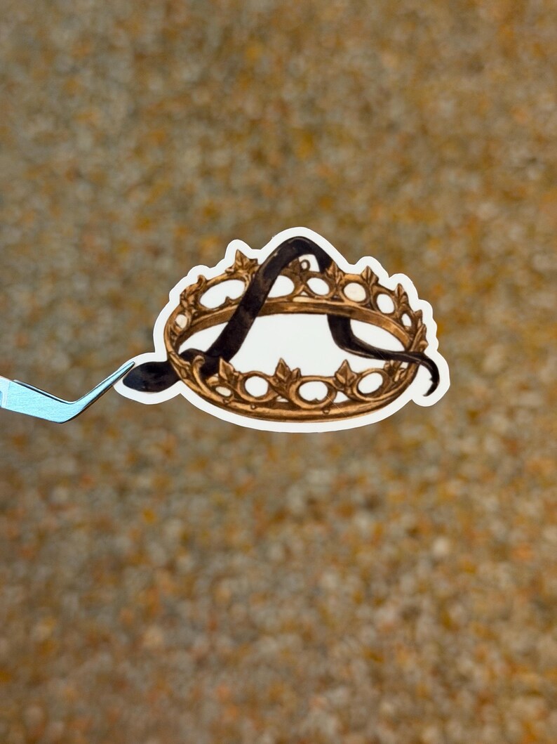 May include: A sticker featuring a gold-colored crown with a black letter "A" inside. The crown has a detailed design with small, circular accents. The sticker is held by a pair of tweezers against a textured, tan background.