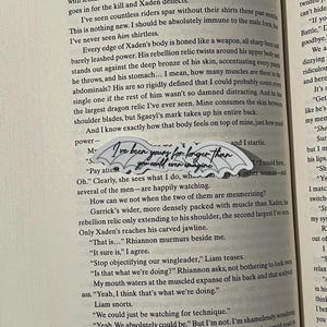 May include: A close-up of a book page with text about a fictional character named Xaden. The text describes Xaden's physique and mentions a dragon relic on his back. The text also includes a quote from a character named Rhiannon, who says "I've been yours for longer than you could even imagine."