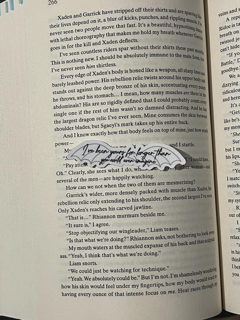 May include: A close-up of a book page with text about a fictional character named Xaden. The text describes Xaden's physique and mentions a dragon relic on his back. The text also includes a quote from a character named Rhiannon, who says "I've been yours for longer than you could even imagine."