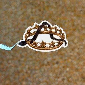 May include: A sticker featuring a gold-colored crown with a black letter "A" inside. The crown has a detailed design with small, rounded shapes. The sticker is held by a pair of tweezers against a textured, tan background.