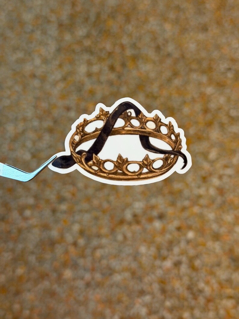 May include: A sticker featuring a gold-colored crown with a black letter "A" inside. The crown has a detailed design with small, rounded shapes. The sticker is held by a pair of tweezers against a textured, tan background.