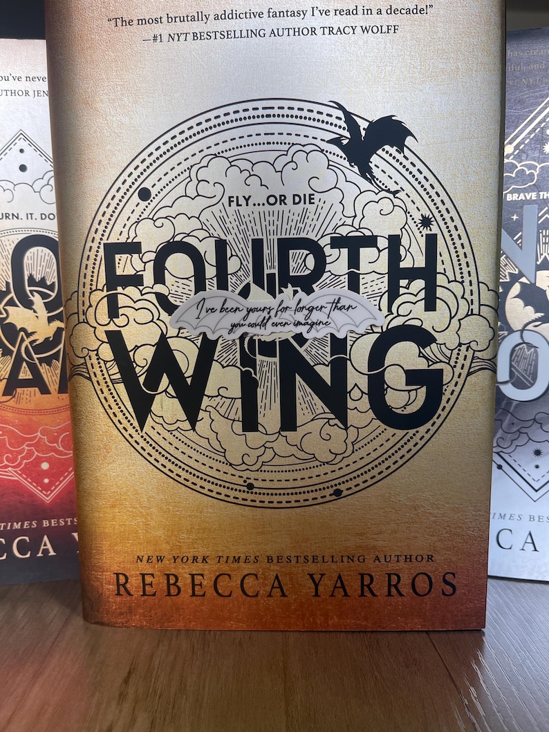 May include: A book cover with a gold background and a black circular design featuring a dragon and the words "Fly...or Die" and "Fourth Wing". The book title is "Fourth Wing" and the author is "Rebecca Yarros".