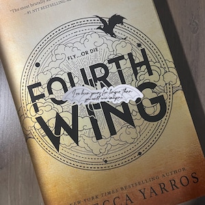 May include: A book cover with a gold background and black text. The title is "Fourth Wing" with the words "Fly...or Die" above it. The author's name is "Rebecca Yarros" and the text "New York Times Bestselling Author" is below it. The book cover features a black dragon with a white wing.