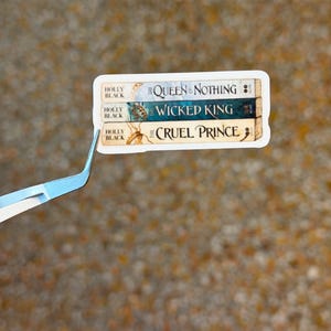 May include: A sticker featuring three book spines: "The Queen of Nothing," "Wicked King," and "Cruel Prince" by Holly Black. The spines are in shades of blue, green, and beige. The sticker is held by a pair of tweezers.
