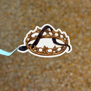 May include: A sticker featuring a gold-colored crown with the letter "A" in black script. The crown has a detailed design with ornate embellishments. The sticker is held by a pair of light blue tweezers against a blurred background.