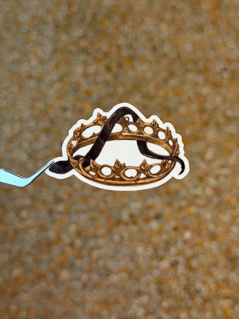 May include: A sticker featuring a gold-colored crown with the letter "A" in black script. The crown has a detailed design with ornate embellishments. The sticker is held by a pair of light blue tweezers against a blurred background.