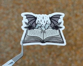 May include: A sticker featuring an open book with mountain peaks and bat wings. The black and white illustration includes roses and stars. The sticker is held by a pair of tweezers.