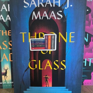 May include: A book cover with a blue and red background featuring a woman holding a sword. The title of the book is "Throne of Glass" by Sarah J. Maas. The spine of the book shows the titles of the series: "Assassin's Blade", "Throne of Glass", "Crown of Midnight", "Heir of Fire", and "Queen of Shadows".