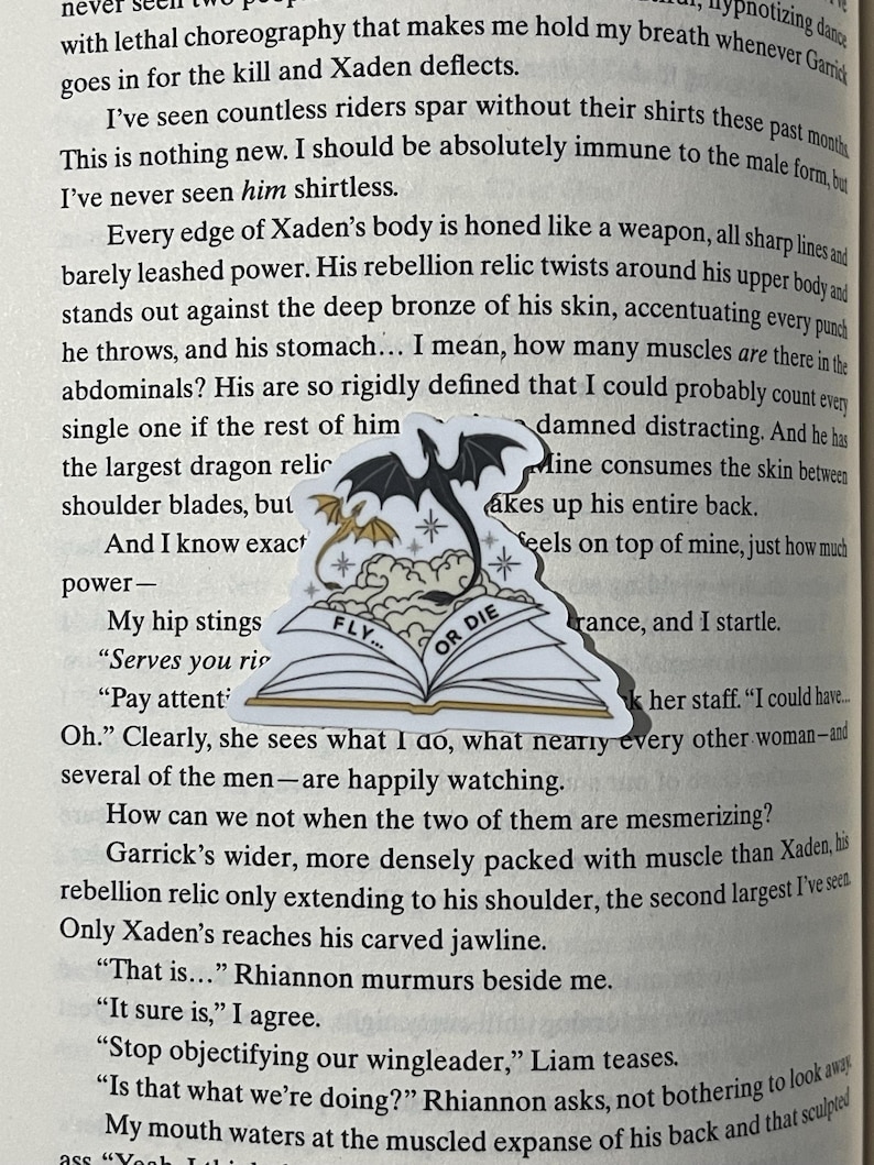 K&ouml;nnte beinhalten: Ein schwarz-wei&szlig;es Buch mit einem Drachen auf dem Cover. Das Buch ist ge&ouml;ffnet und die Seiten sind sichtbar. Das Buch tr&auml;gt die Worte "Fly... or Die" auf dem Cover. Das Buch liegt auf einer Seite eines Buches mit Text &uuml;ber einen Charakter namens Xaden.