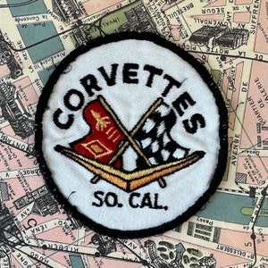 May include: A white, round patch with black stitching around the edges. The patch features a black and white checkered flag and a red flag with a yellow star crossed over each other. The text "CORVETTES SO. CAL." is printed in black on the patch.