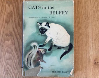 Może przedstawiać: Książka w twardej oprawie zatytułowana CATS in the BELFRY Reminiscences of a Siamese Cat Lover autorstwa Doreen Tovey. Okładka ma jasnoniebieskie tło z ilustracją kota syjamskiego i wiewiórki. Dolna krawędź okładki jest podarta.