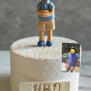 May include: A white cake with a fondant figurine of a boy wearing a blue shirt and khaki shorts. The cake is decorated with a white plaque that says "HBD Raghav".