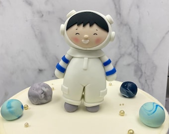 May include: A white fondant cake decorated with a blue and white astronaut figurine, blue and white planets, and gold sprinkles.