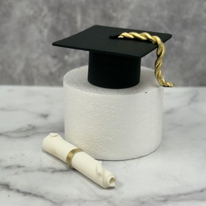 Op de afbeelding: Een zwarte graduatiecap met een gouden kwast staat op een witte taartstandaard. Voor de cap ligt een witte rol met een gouden lint.