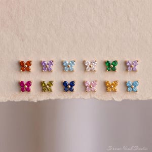 May include: A collection of twelve small butterfly-shaped stud earrings arranged in two rows. Each earring features four teardrop-shaped gemstones in various colors including red, purple, light blue, white, green, pink, yellow, and dark blue.