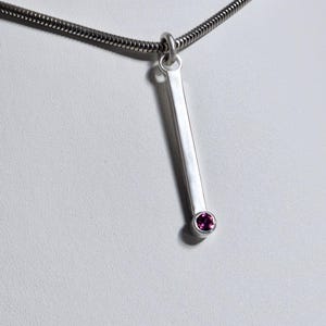 May include: A silver-toned necklace featuring a smooth, vertical rectangular bar pendant. At the base of the bar, a single, small, round, deep pink gemstone is set in a bezel. The pendant hangs from a thin, flexible silver-toned snake chain.