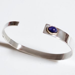 May include: A silver cuff bracelet with a rectangular gold accent holding a dark blue gemstone. The open-ended bracelet is approximately 2.5 inches in diameter. The jewelry is set against a white background.
