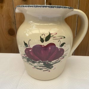 May include: Cream-colored ceramic pitcher with a large handle and a spout. The pitcher is decorated with a cluster of purple cherries and green leaves. The rim has a blue and white scalloped design.