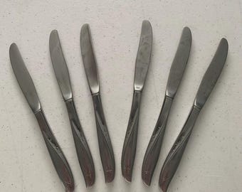 May include: Set of six stainless steel butter knives. Each knife has a curved handle with a decorative starburst design. The blades are smooth and rounded. The knives are arranged on a light-colored surface.