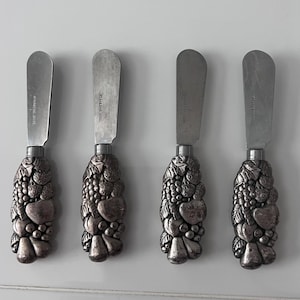 May include: A set of four butter knives with silver-toned metal handles featuring a detailed, raised relief design of various fruits including grapes, pears, and strawberries. Each blade is made of stainless steel with the text STAINLESS STEEL.