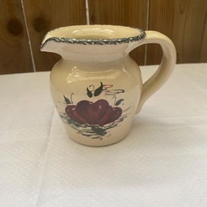 May include: Cream-colored ceramic pitcher with a handle and spout. The pitcher features a hand-painted design of red apples with green leaves. The rim is decorated with a dark blue and green pattern. A decorative kitchenware item.
