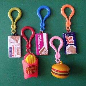 May include: Five colorful plastic keychains with different candy bar and food themed charms. The charms include a chocolate bar, a milk chocolate bar, a french fries container, a hamburger, and a candy bar with the text "Cadbury's Bar Six".