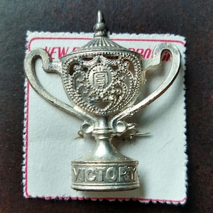 May include: A silver-colored trophy pin with a detailed design, including handles and a lid. The word "VICTORY" is engraved on the base. The pin is on a white card with red trim, against a brown background.