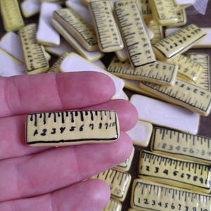 May include: A collection of small, rectangular, yellow and white ceramic rulers. The rulers are marked with black lines and numbers from 1 to 10. One ruler is held in a hand, with others scattered around.