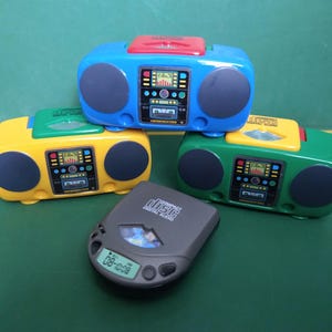 May include: Three toy boomboxes in blue, yellow, and green, stacked on top of each other. A gray toy CD player with the text "Compact Disc Digital Audio" is in front of the boomboxes.