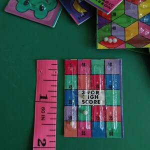 May include: A close-up of a game board with a clear plastic grid and colorful squares. The board has numbers printed on it, including 5, 10, 15, 20, 25, 30, 35, 40, 45, 50, 55, and 60. The text "3 for High Score" is printed in the center of the board. There are three white marbles in the grid.