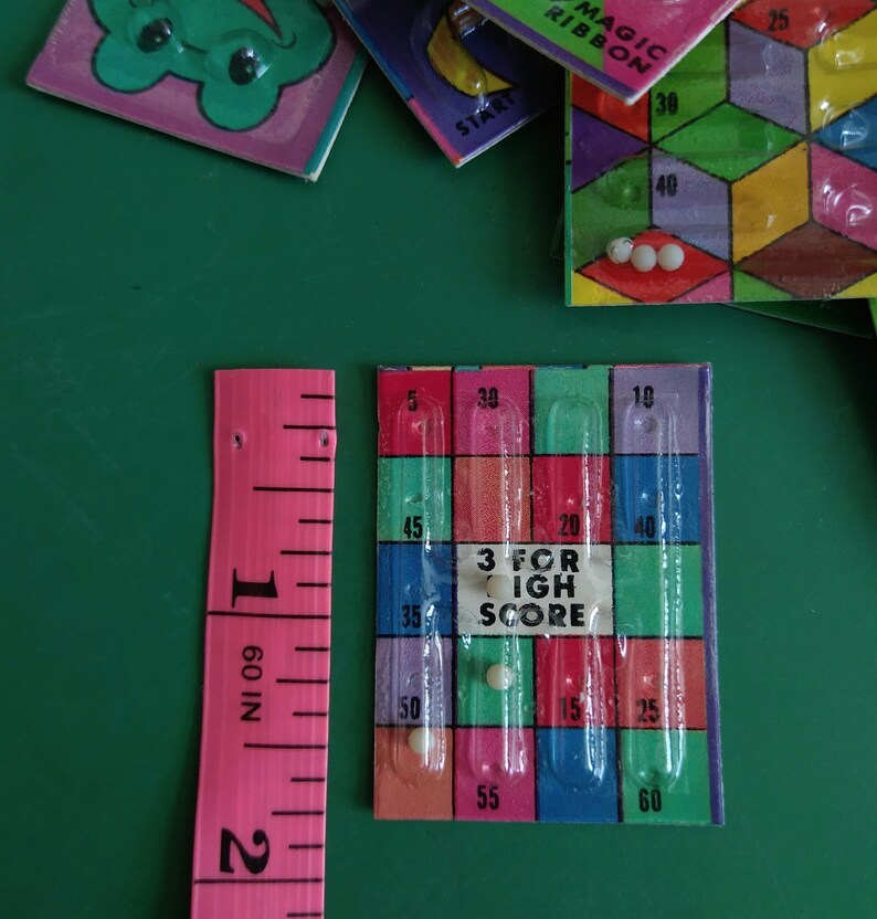May include: A close-up of a game board with a clear plastic grid and colorful squares. The board has numbers printed on it, including 5, 10, 15, 20, 25, 30, 35, 40, 45, 50, 55, and 60. The text "3 for High Score" is printed in the center of the board. There are three white marbles in the grid.