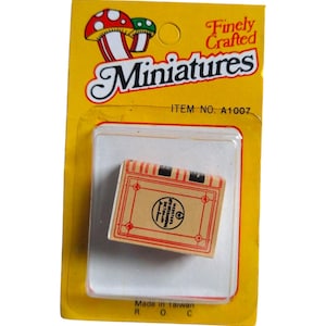 May include: A miniature dictionary packaged on a yellow card with the text "Finely Crafted Miniatures." The dictionary is rectangular with a red border and the text "WEBSTERS DICTIONARY." The package reads "Made in Taiwan."