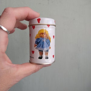 May include: White metal tin with a red heart pattern. The tin features a picture of a doll with blonde hair wearing a blue dress and white shoes. The text on the tin reads "Sweets LTD."
