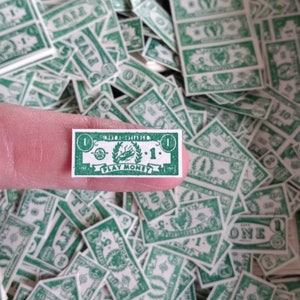 May include: A close-up of a single green play money bill with the number "1" and the words "PLAY MONEY" printed on it. The bill is held by a finger against a background of many other green play money bills.