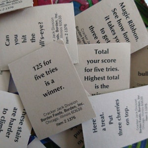 May include: A close-up of several white cards with black text. The cards are from a Cracker Jack game and have instructions for playing the game. The text on the cards includes "125 for five tries is a winner", "Total your score for five tries. Highest total is the winner", "Can you hit the bull's-eye?", and "Here's a treat. Put three cherries on top."