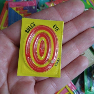 May include: A yellow and red Bull's Eye maze game with a white marble in the start position. The game is held in a hand.