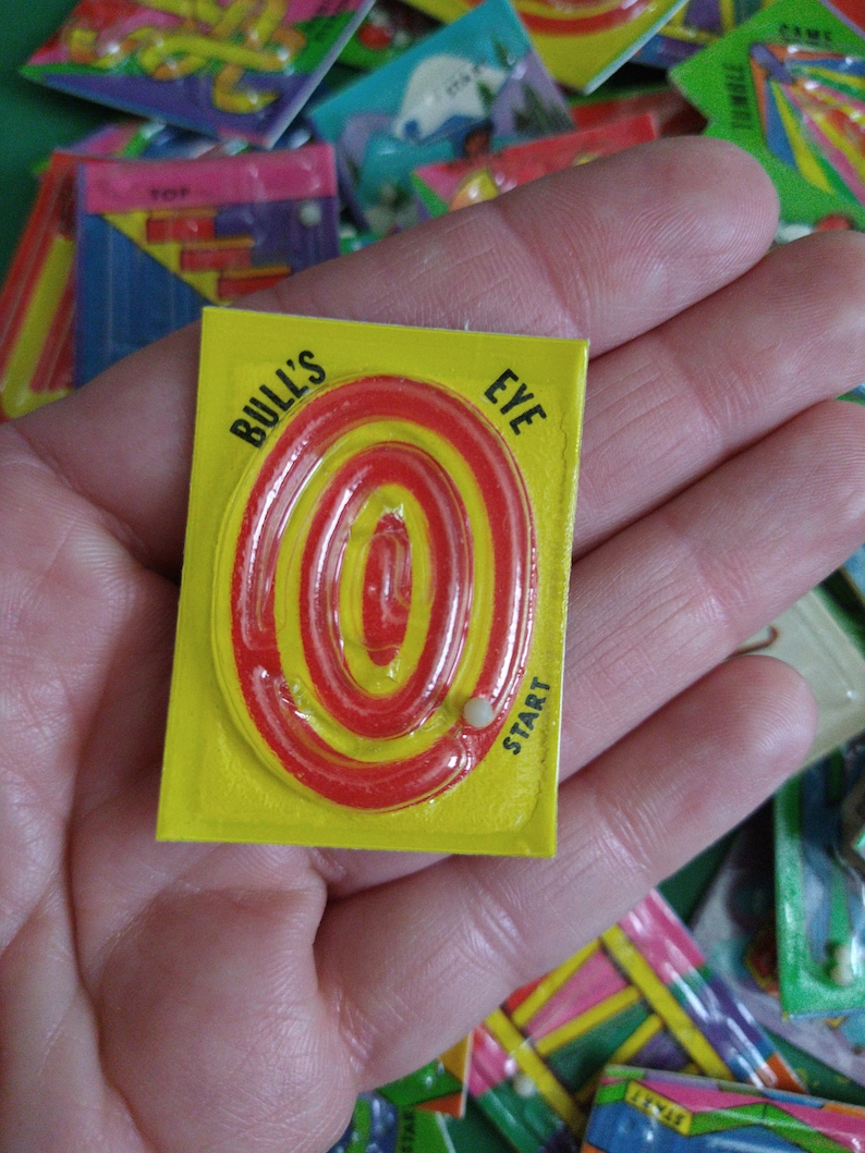 May include: A yellow and red Bull's Eye maze game with a white marble in the start position. The game is held in a hand.