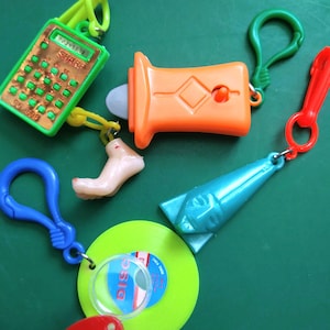 May include: A collection of colorful plastic keychain toys, including a calculator, a whistle, a foot, a pyramid, and a green disc with a magnifying glass. The toys are all attached to metal rings and have a variety of colors, including green, orange, blue, and red.