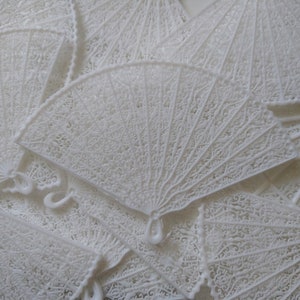 May include: A pile of white, intricately designed, paper folding fans. The fans are made of delicate, lacy material and are arranged in a random pattern.