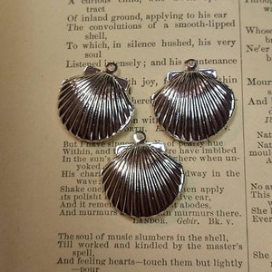 May include: Three shiny silver seashell charms with a textured surface, each with a small loop at the top for attaching to jewelry. The charms are displayed on a vintage book page with the word "SHELL" at the top.