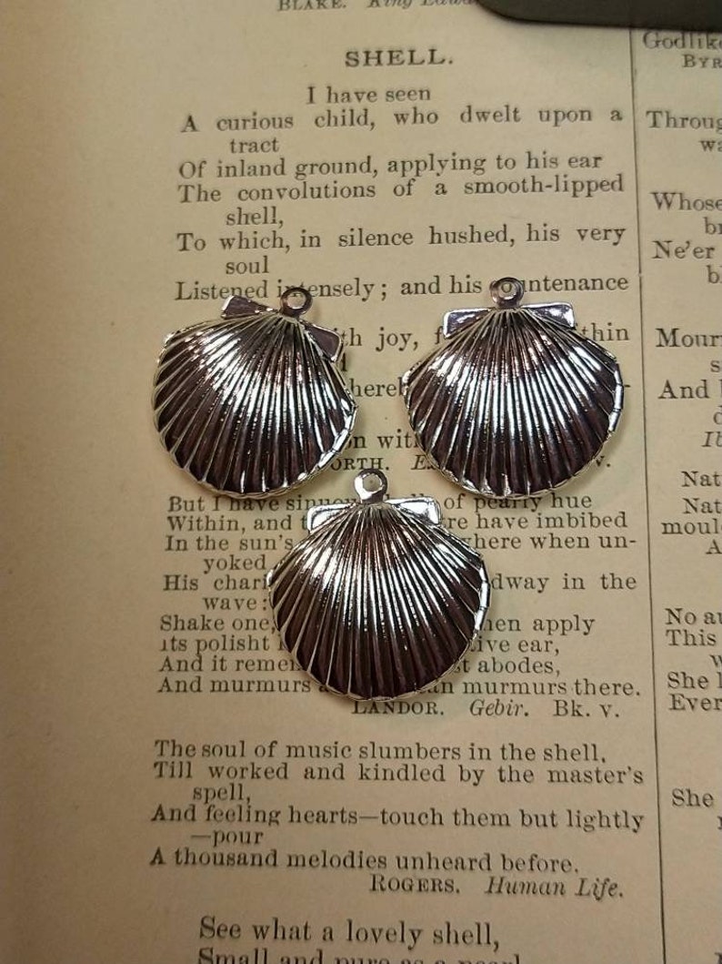 May include: Three shiny silver seashell charms with a textured surface, each with a small loop at the top for attaching to jewelry. The charms are displayed on a vintage book page with the word "SHELL" at the top.