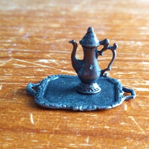 May include: A miniature silver teapot with a decorative handle sits on a silver tray with a scalloped edge.