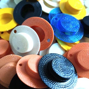 May include: A collection of colorful plastic hat-shaped buttons with various textures and designs. The buttons are in shades of yellow, orange, blue, black, and white.