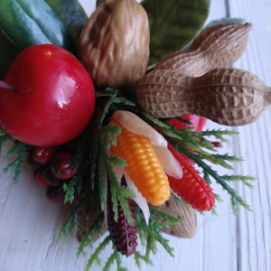 May include: A close-up of a fall-themed artificial fruit and nut arrangement. The arrangement features a red apple, a brown nut, a peanut, a yellow and orange corn cob, and a red and white corn cob. The arrangement is surrounded by green leaves.