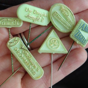 May include: A collection of six vintage green plastic advertising pins. The pins feature the text "Verdun's", "Uw Drogist heeft it", "Droppinia", "Pri", "ANIMO ZEGELS".
