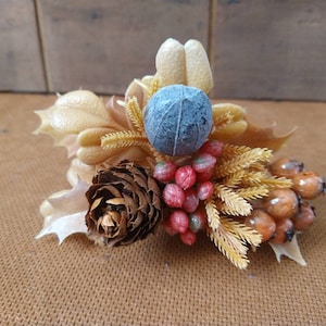 May include: A decorative floral arrangement featuring a small pine cone, blue sphere, and various berries in shades of red and orange. The arrangement includes beige and tan leaves and sprigs, creating a natural, autumnal aesthetic.