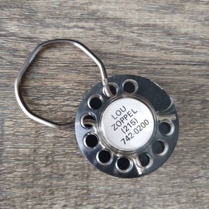 May include: A silver metal bottle stopper with a round, circular design. The stopper has a ring handle and a white label with the text "LOU ZOPPEL (215) 742-0200".