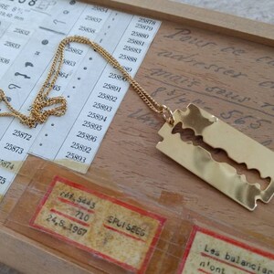 May include: A gold chain necklace with a pendant shaped like a razor blade. The pendant is hanging on a wooden surface with writing on it.