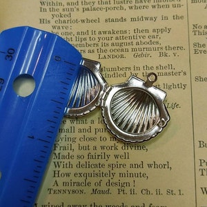 May include: Open silver seashell pendant, approximately 1 inch in diameter, with a hinged design. The pendant is open, revealing a detailed interior. A blue ruler is placed next to the pendant, and the background is a vintage book page.