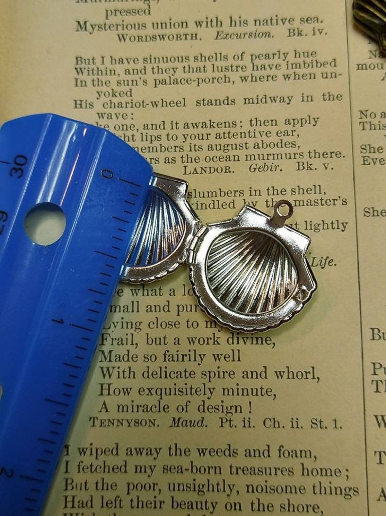 May include: Open silver seashell pendant, approximately 1 inch in diameter, with a hinged design. The pendant is open, revealing a detailed interior. A blue ruler is placed next to the pendant, and the background is a vintage book page.